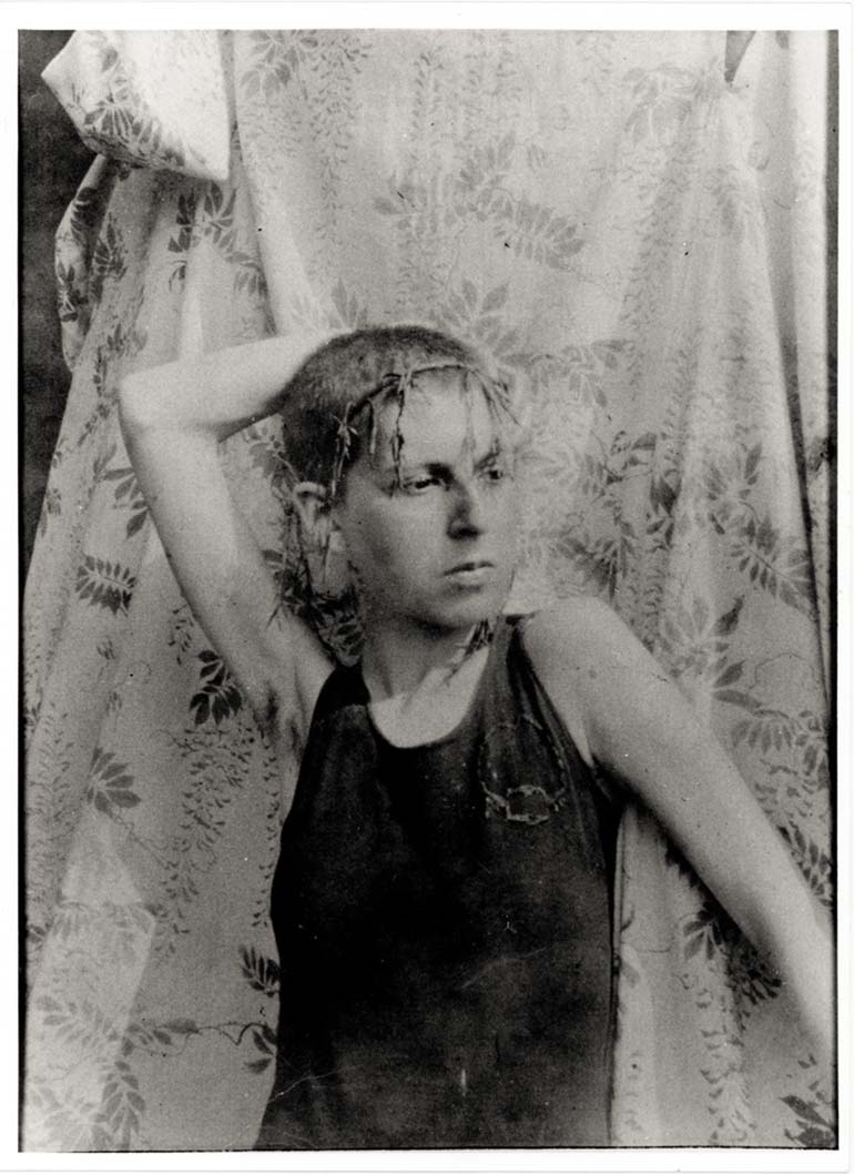 Claude Cahun :: Autoportrait, vers 1919. Autoportrait de Claude Cahun jeune fille. Les cheveux courts, elle arbore une couronne de feuilles nouées en fines tresses et pose devant un tissu clair à motifs végétaux. Tirage argentique sépia. | Musée d'arts de Nantes
