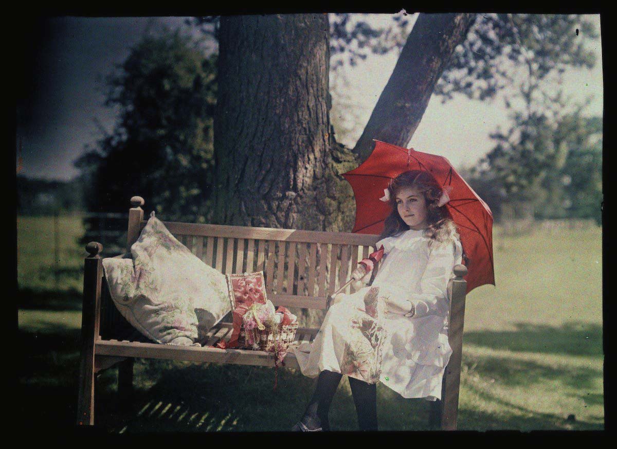 An autochrome of her daughter sitting under a tree, taken by Etheldreda Janet Laing. Next to her, a basket overflows with ribbons and thread as she holds a length of fabric she may be embroidering. She carries a parasol to shade her face from the bright sunshine.