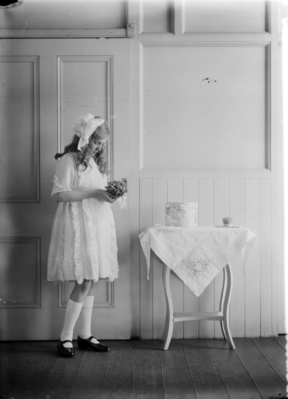Image shows a girl, whole-length, standing beside table, holding a bouquet of flowers.